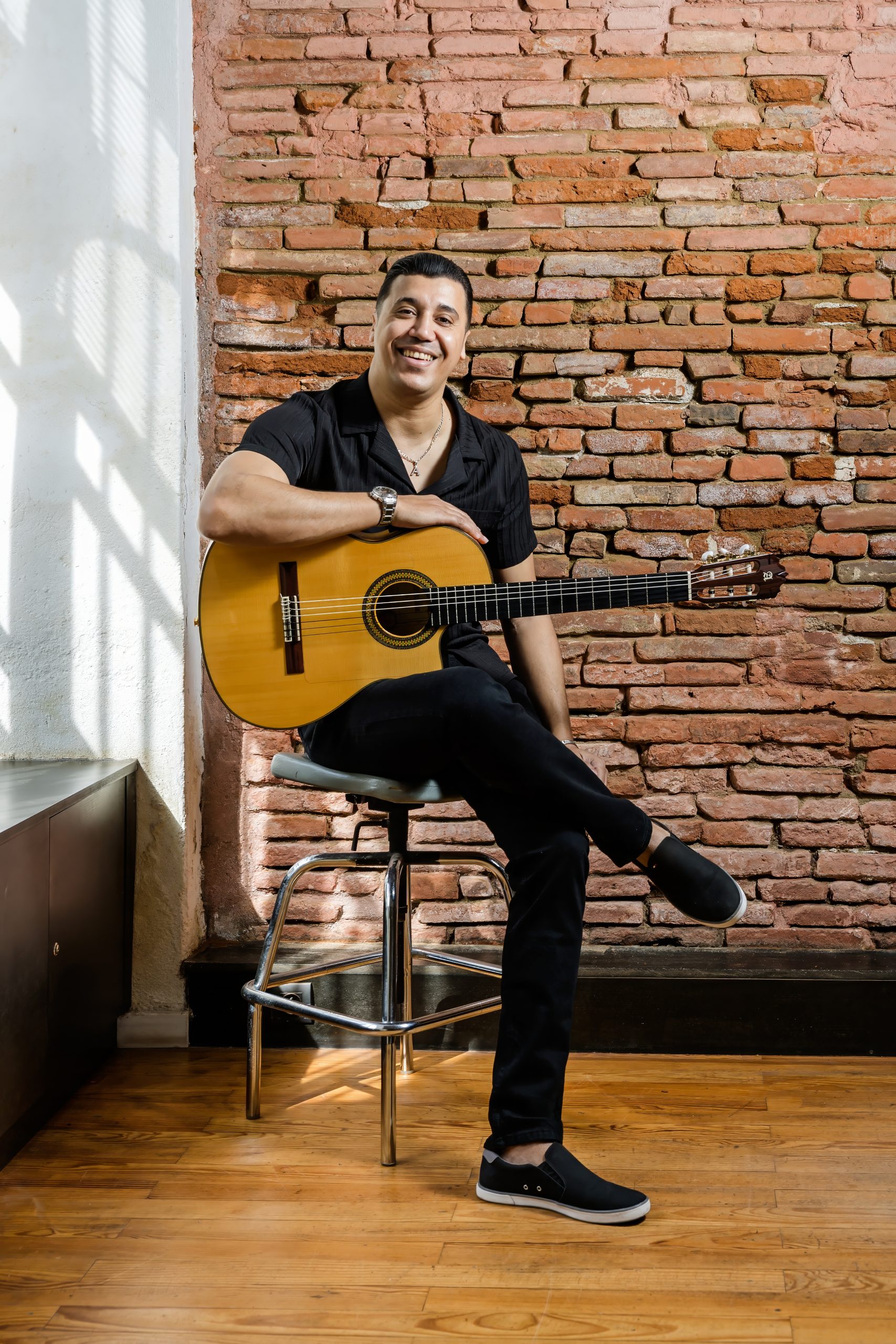 Amine Belayachi assis devant un mur de briques avec sa guitare